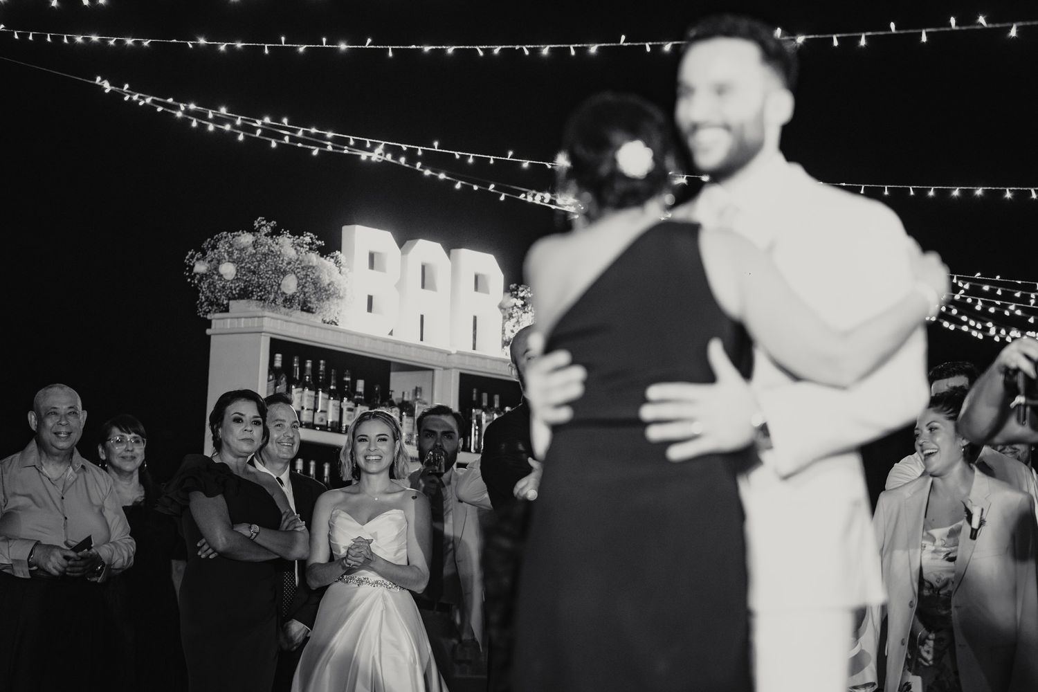 Couple sharing their first dance at an outdoor wedding reception with string lights and a bar backdrop.