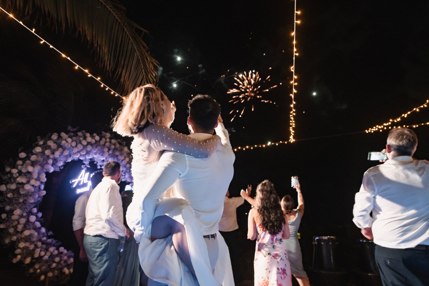 Groom lifts bride joyfully as fireworks light the night sky during outdoor wedding reception.