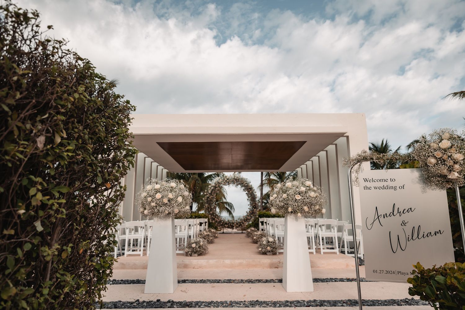 Elegant outdoor wedding ceremony setup with floral arch, white chairs, and welcome sign for Andrea and William.