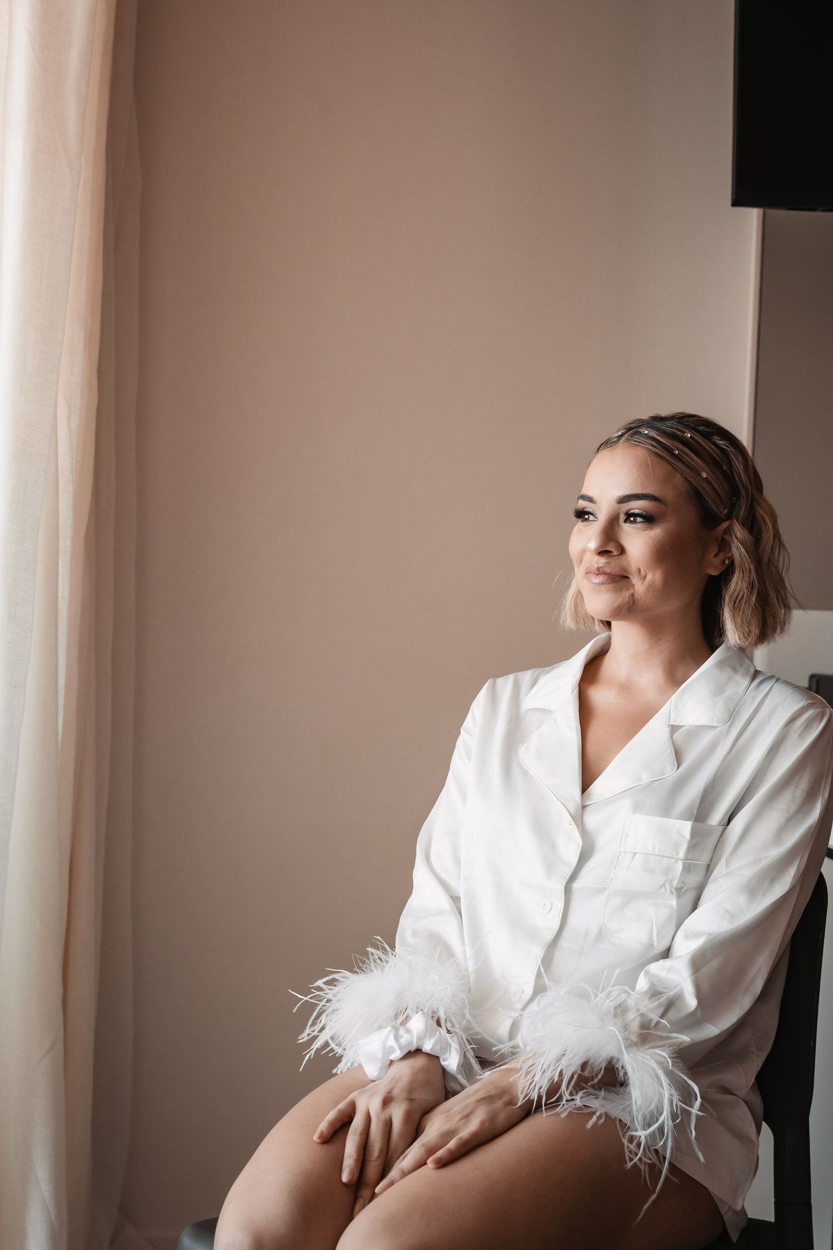 Bride in white feather-trimmed satin pajamas sitting gracefully by a window in soft natural light.