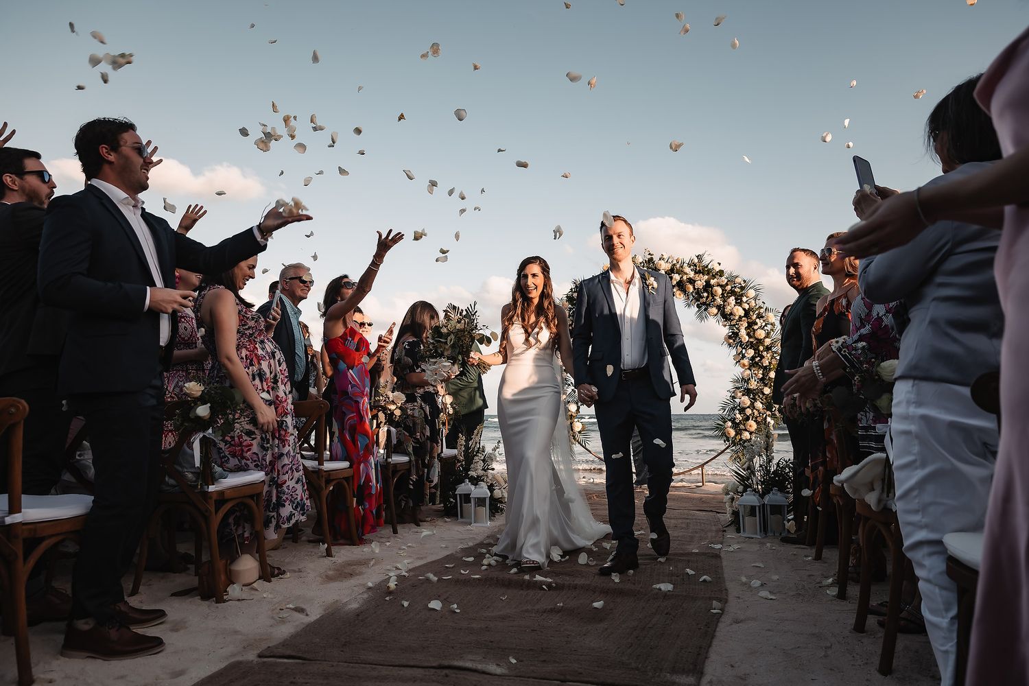 Bride and groom walk down outdoor aisle as guests toss white flower confetti at a romantic seaside wedding ceremony.