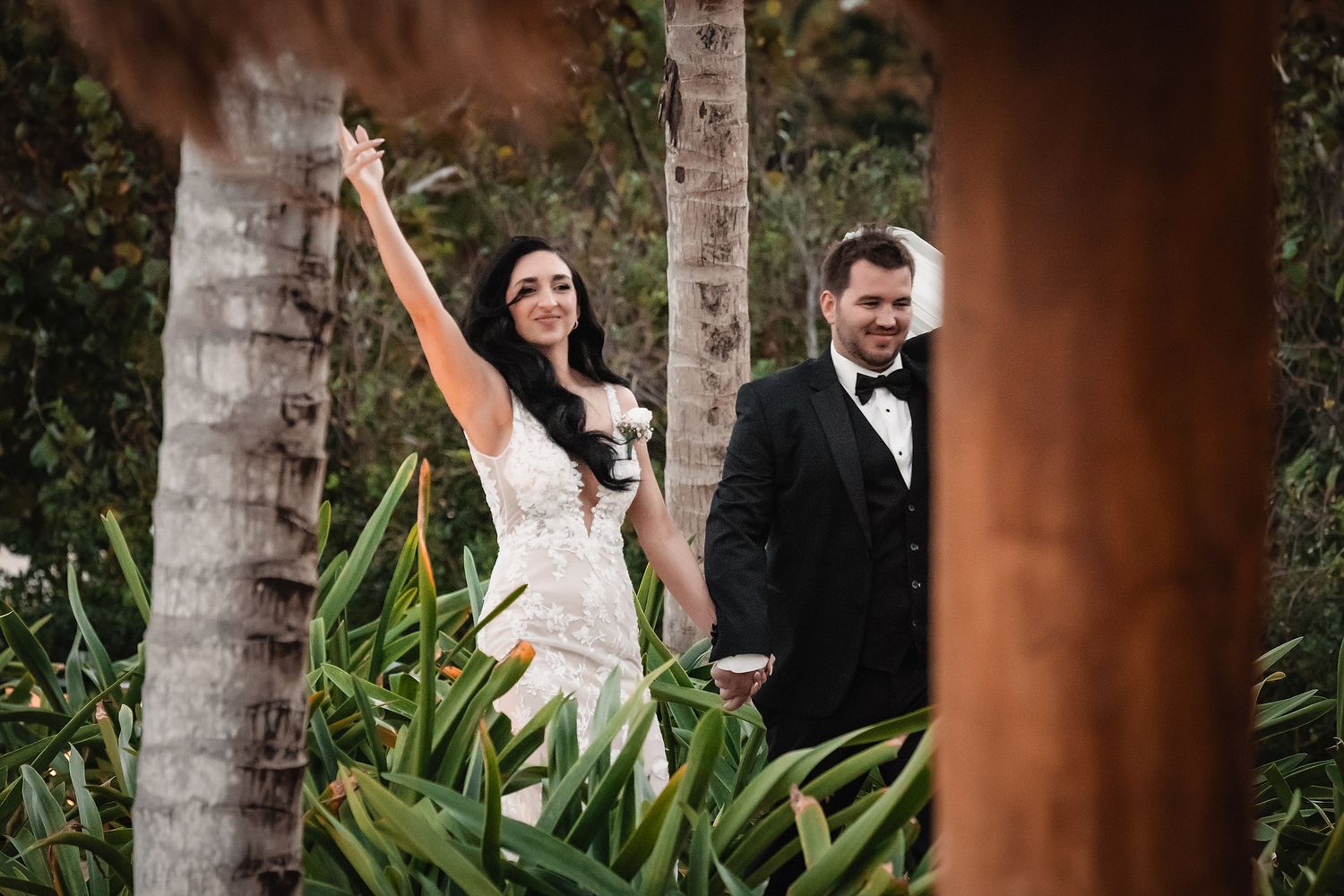Joyful bride in white gown raises arm among tropical palms as groom in tuxedo smiles beside her.