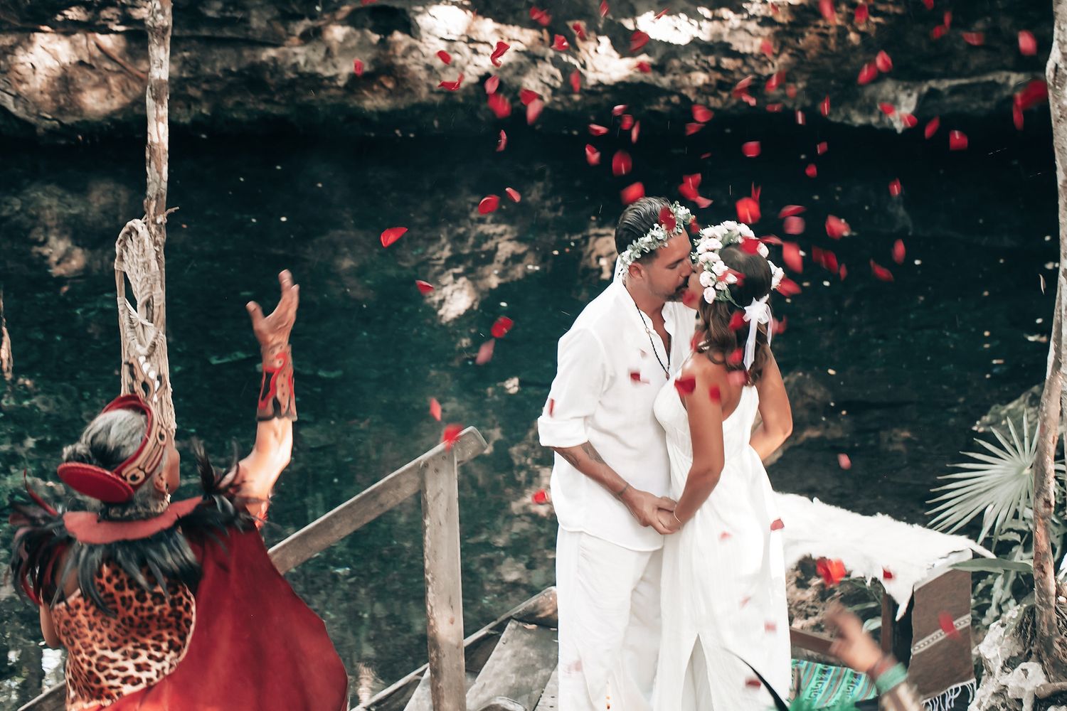 A bride and groom embrace and kiss surrounded by flower petals in a lush outdoor setting.