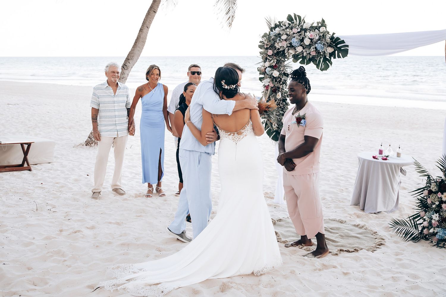 Image of a bride and groom embracing on a beach with other wedding guests.