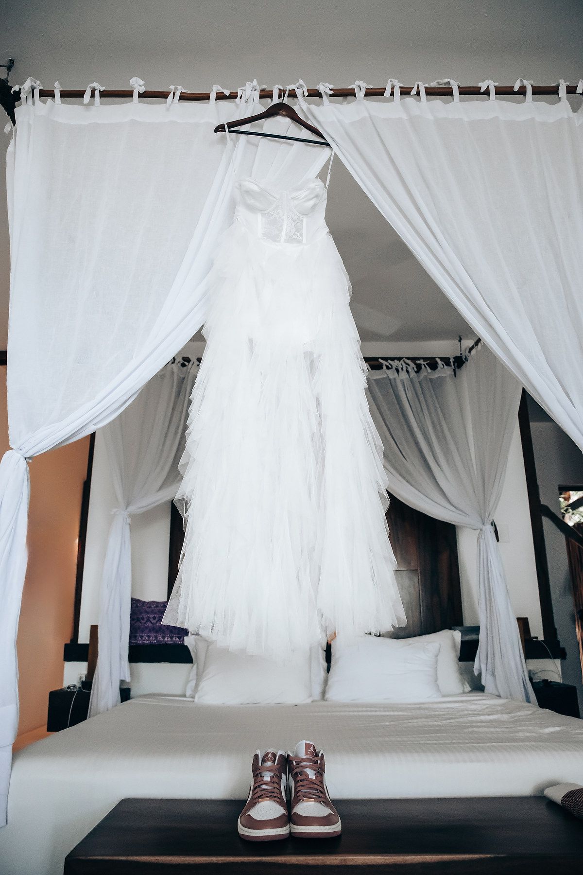 A white wedding dress hangs on a clothing rack in a room with white curtains.