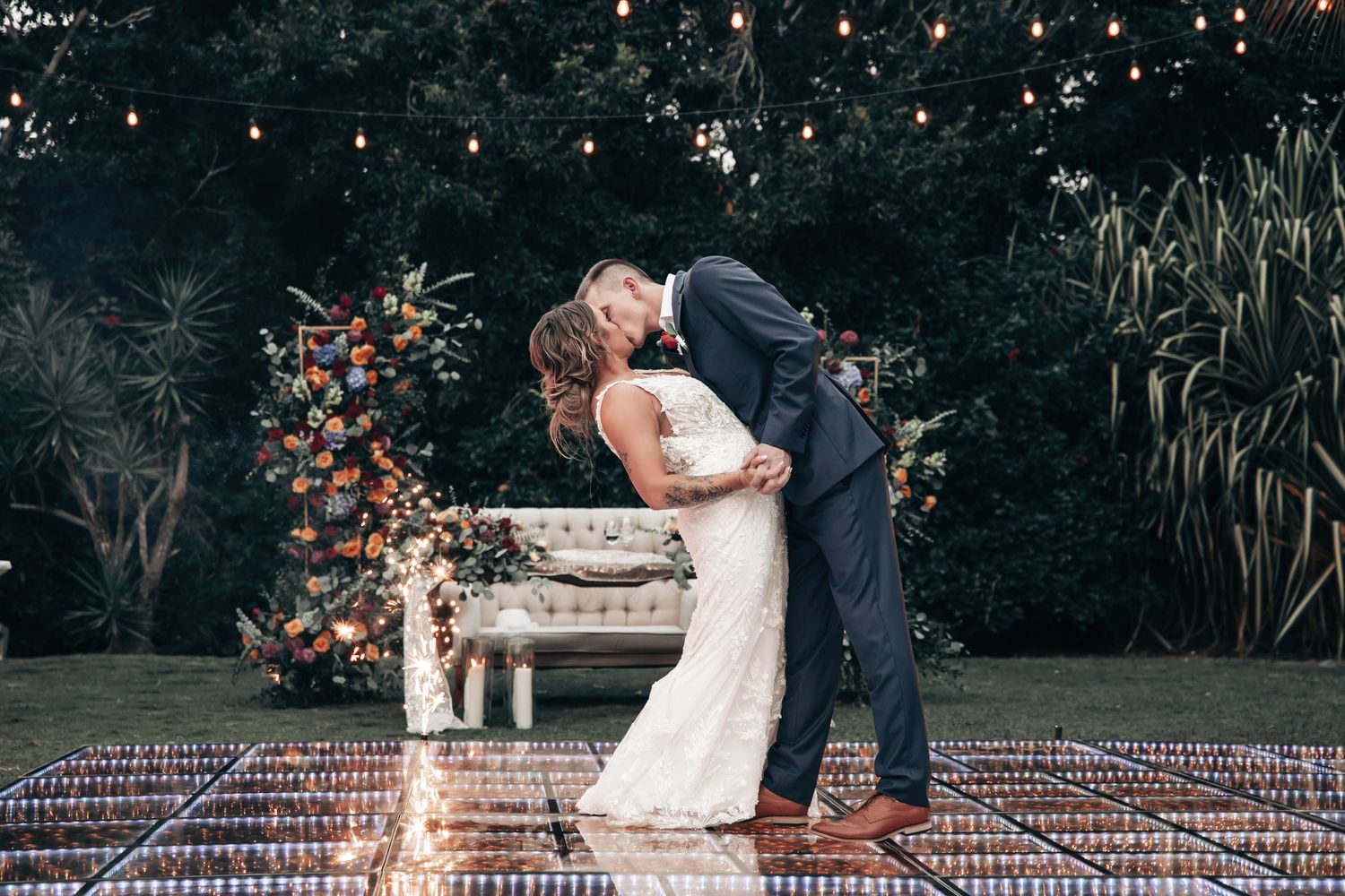 Bride and groom sharing first kiss on illuminated dance floor with floral backdrop and string lights.