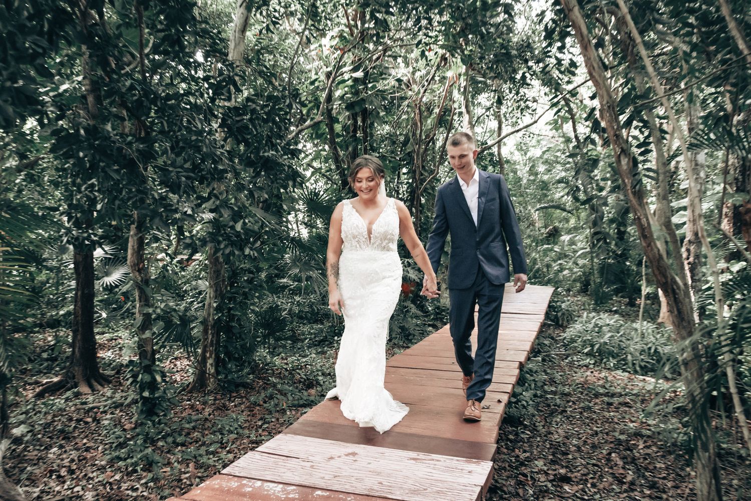 Bride and groom walking hand in hand along a wooden boardwalk path through a beautiful tropical forest setting.