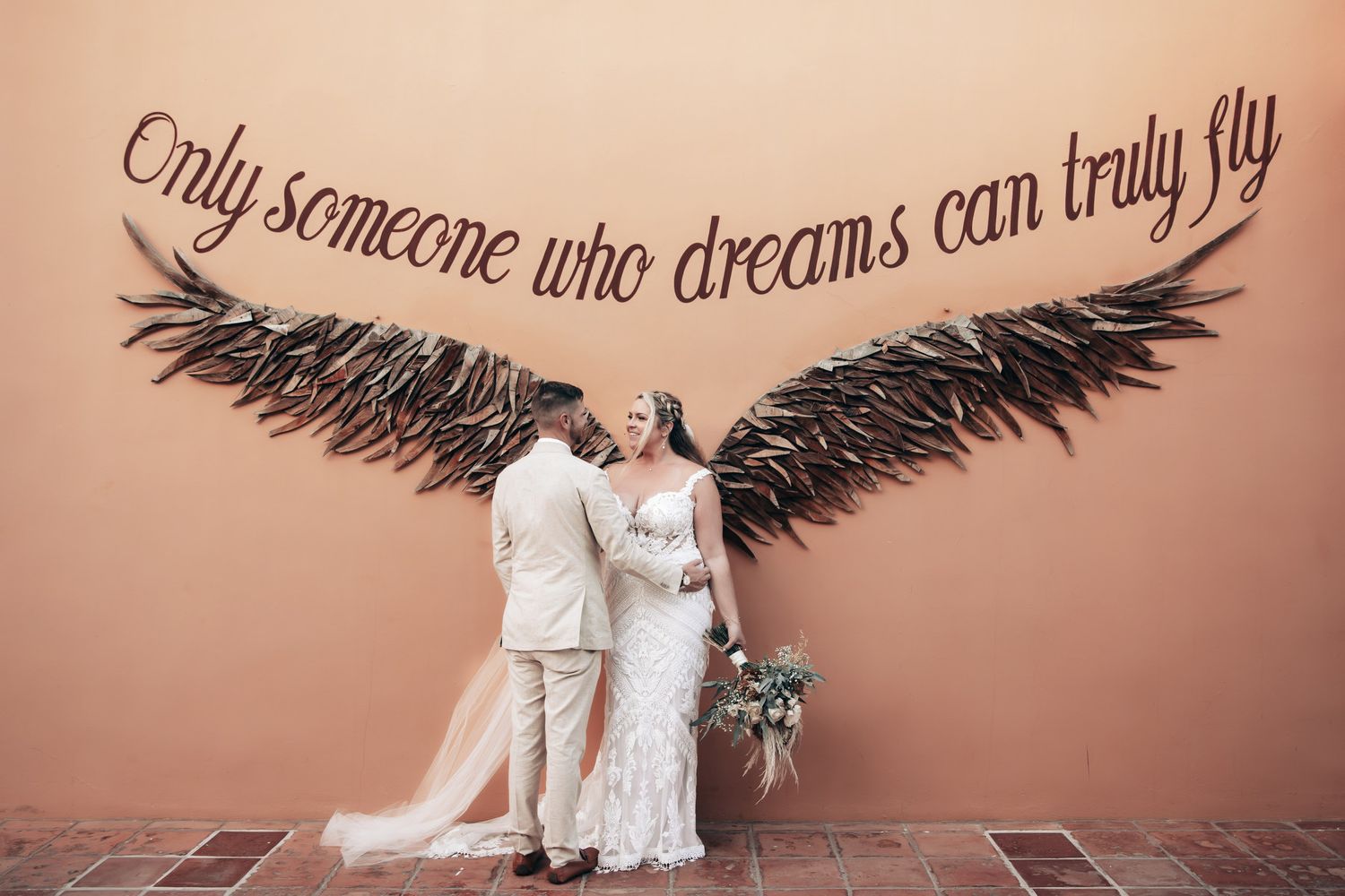 Newlyweds kissing beneath large angel wings wall art with inspirational quote at wedding photo location at Dreams Tulum