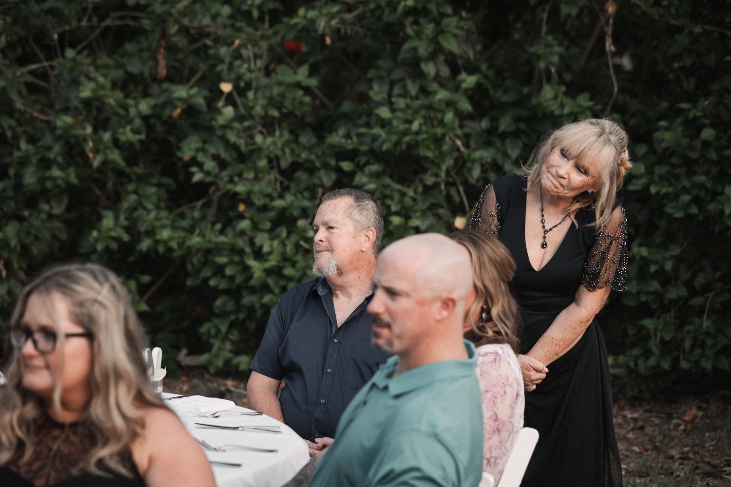 Wedding guests in elegant black attire gather outdoors at a beautifully set reception dinner table.