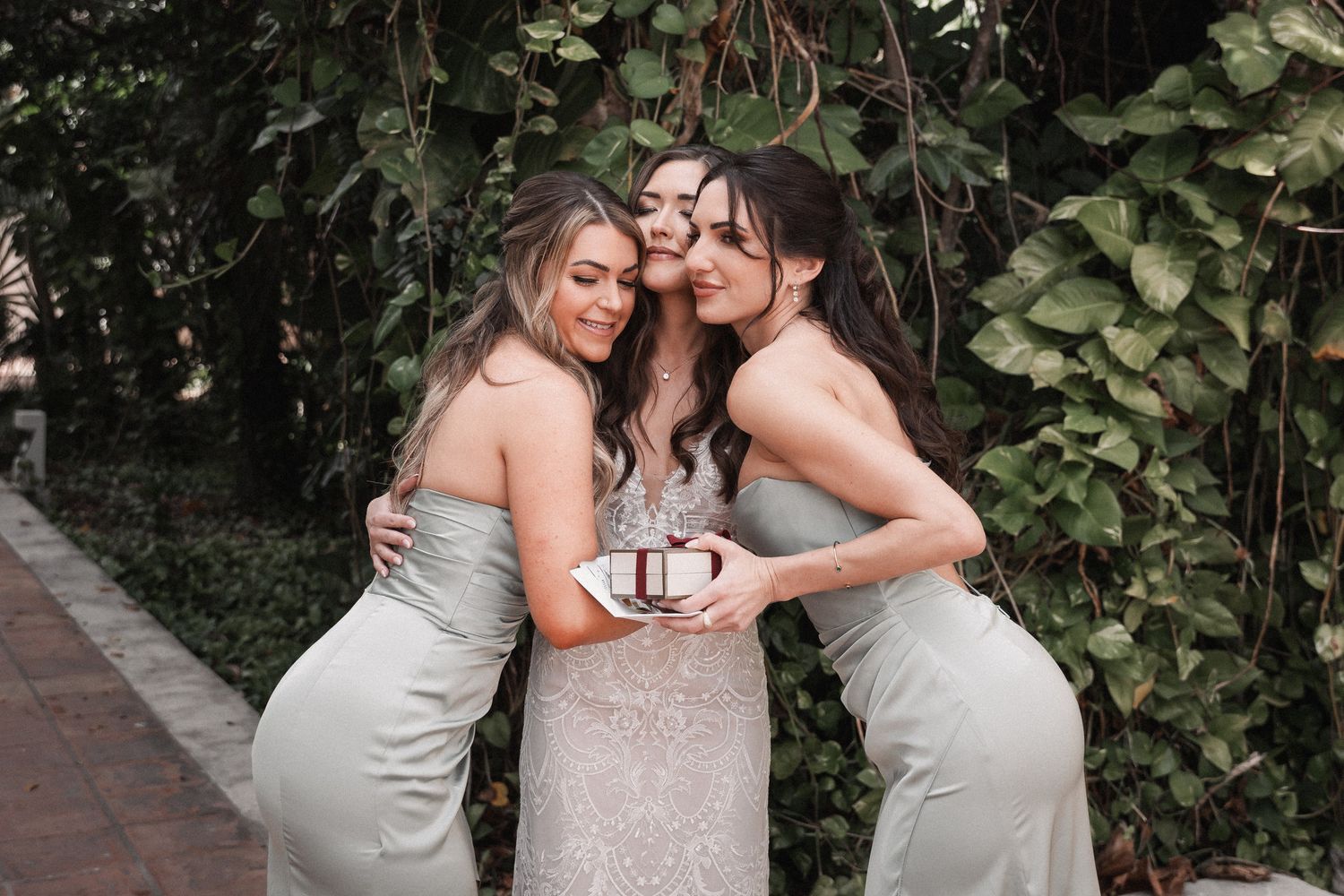 Bride in lace gown embraced by two bridesmaids in sage green dresses holding white bouquets in garden setting.