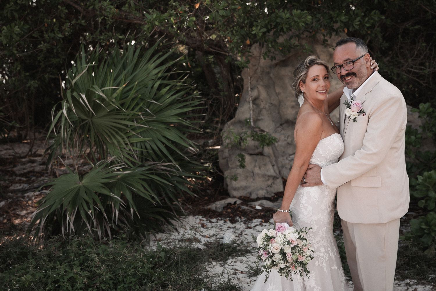 Bride and groom posing together in tropical garden setting with lush greenery and stone backdrop.