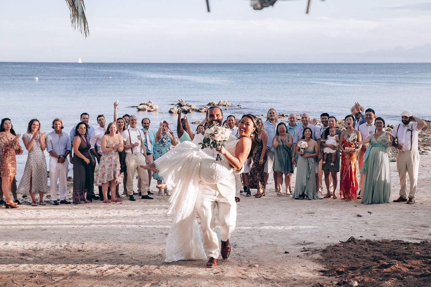 Beachfront wedding celebration with guests gathered on sandy shore under blue skies.