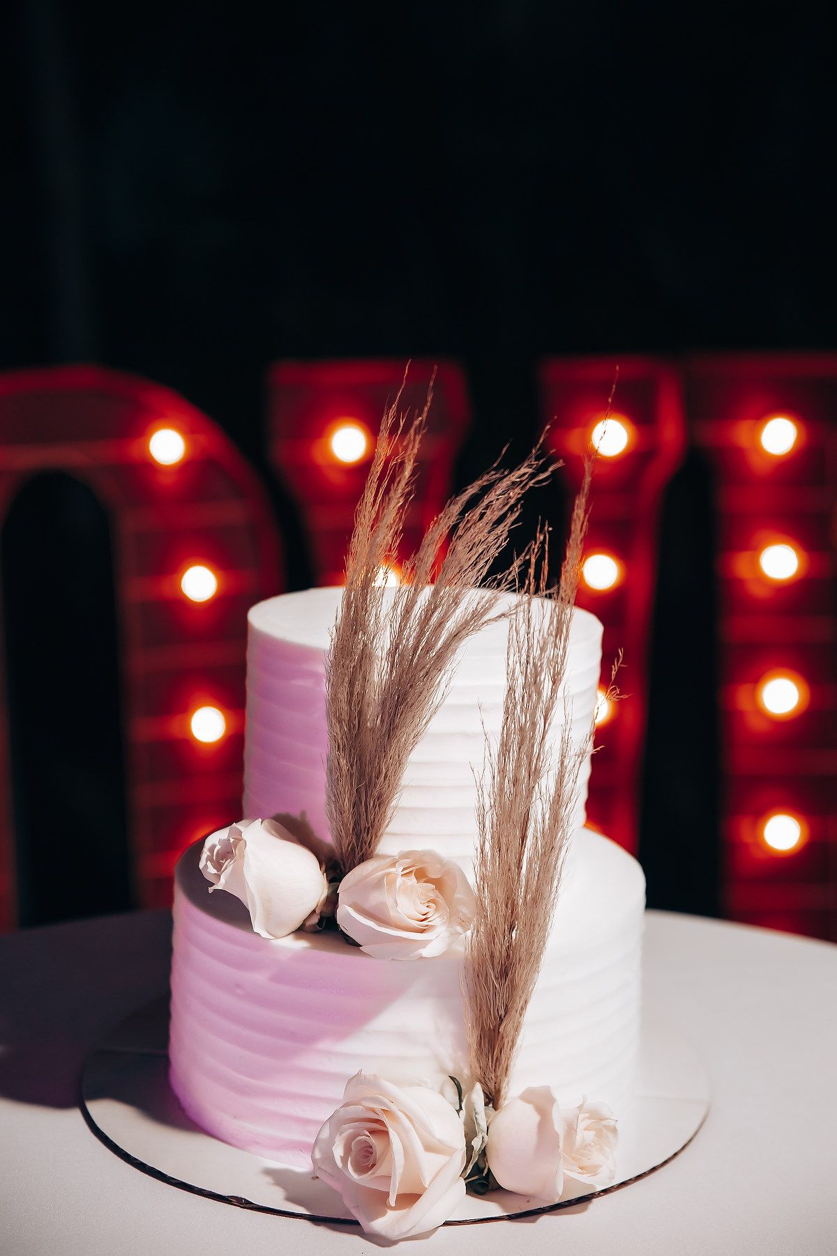 White wedding cake with blush roses and wheat stalks against red light backdrop.