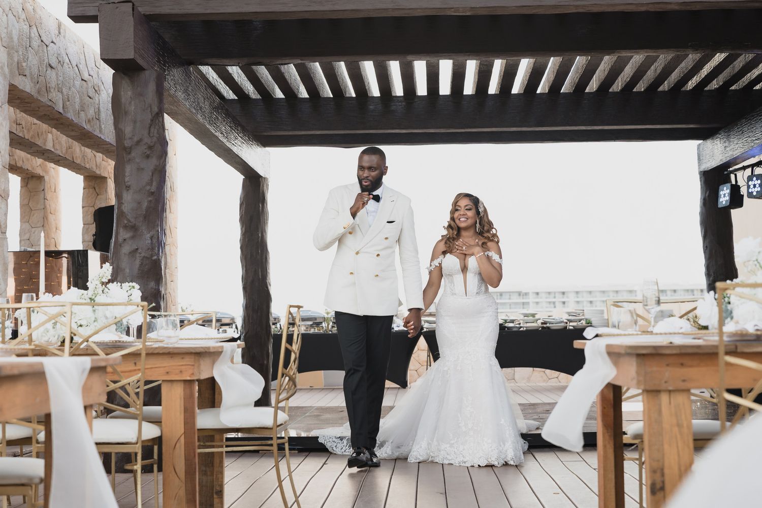 Bride and groom in matching white attire standing together on a decorated outdoor reception venue deck.