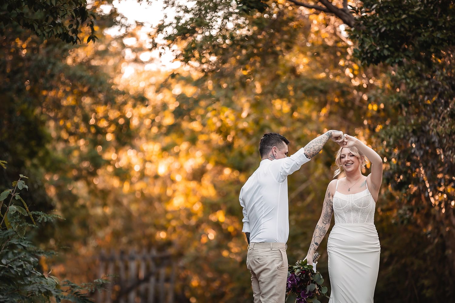 Groom twirling bride in white dress among golden autumn trees in a romantic outdoor wedding photo.