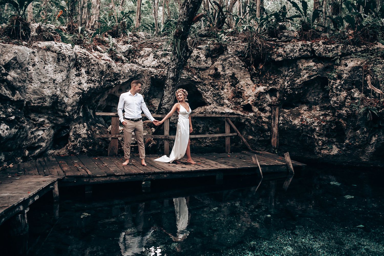 Two people in white outfits stand on a wooden platform beside a dark cenote pool surrounded by rocky walls in Mexico.