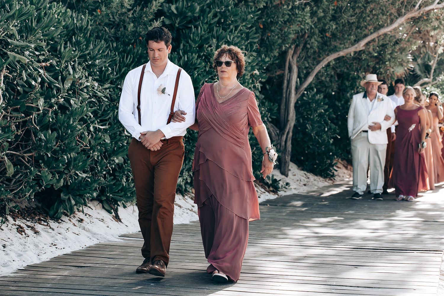 Wedding guests walk along a scenic outdoor pathway lined with trees and white decor.