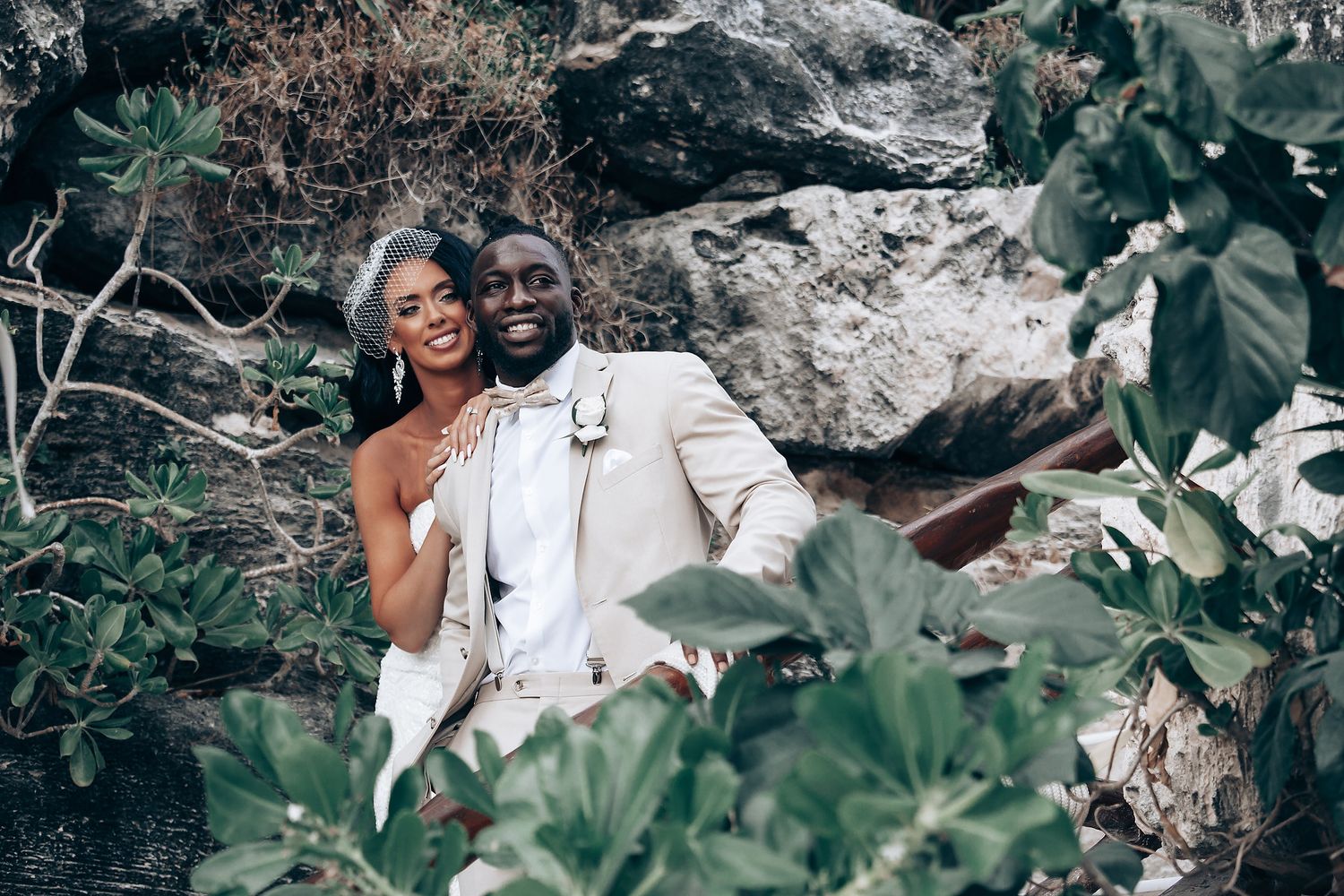 A romantic couple poses together outdoors among lush greenery and rocks in a natural setting.