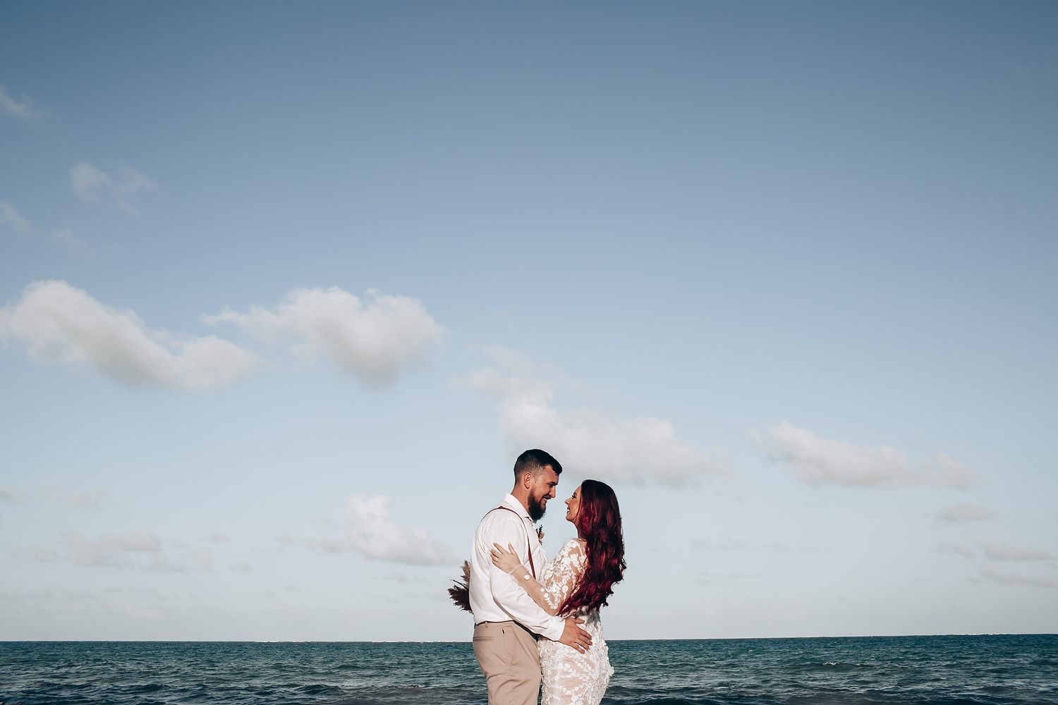 Romantic couple embracing on beach with ocean and blue sky backdrop during destination wedding.