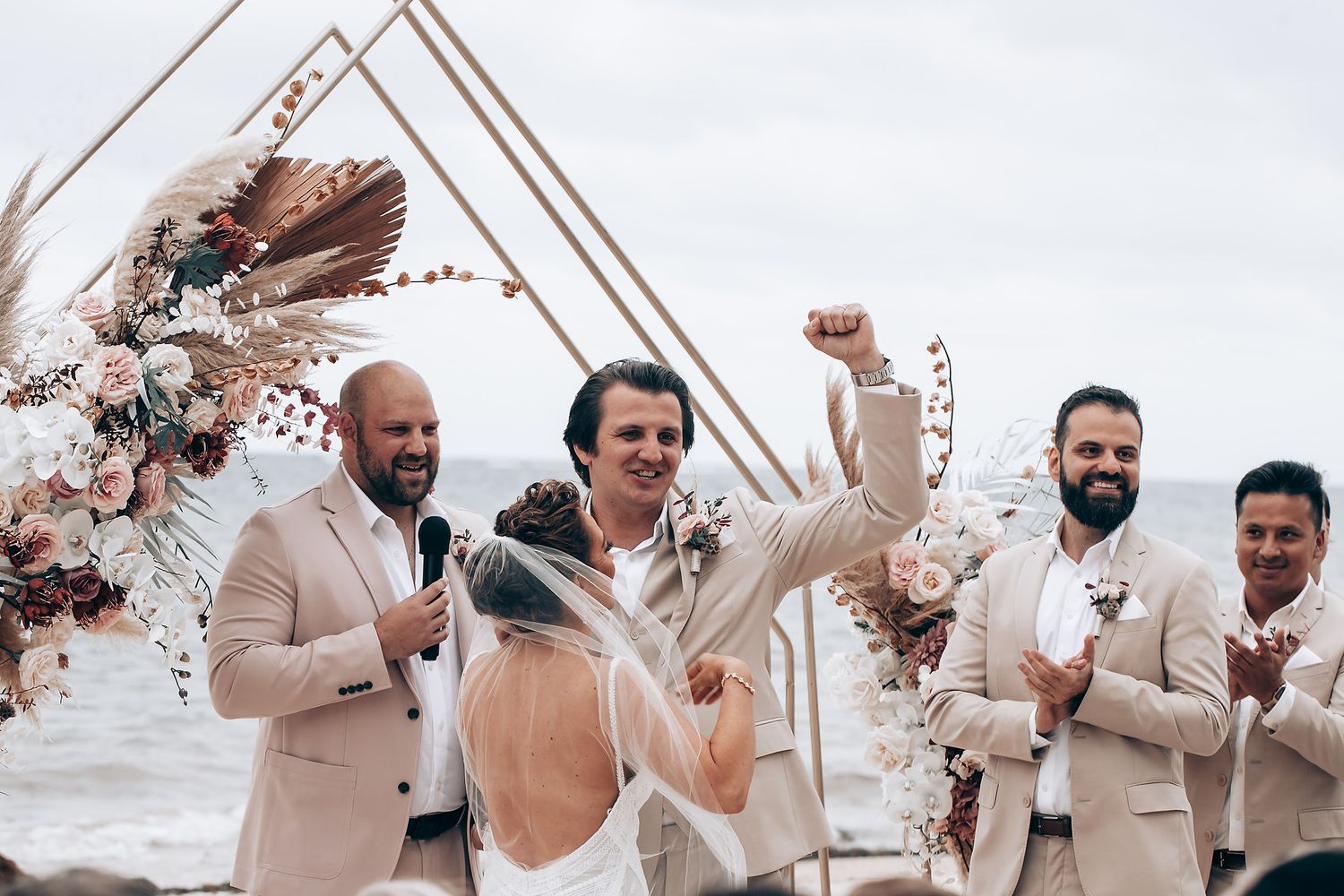 Wedding ceremony celebration with guests in beige attire cheering under a geometric arch frame.