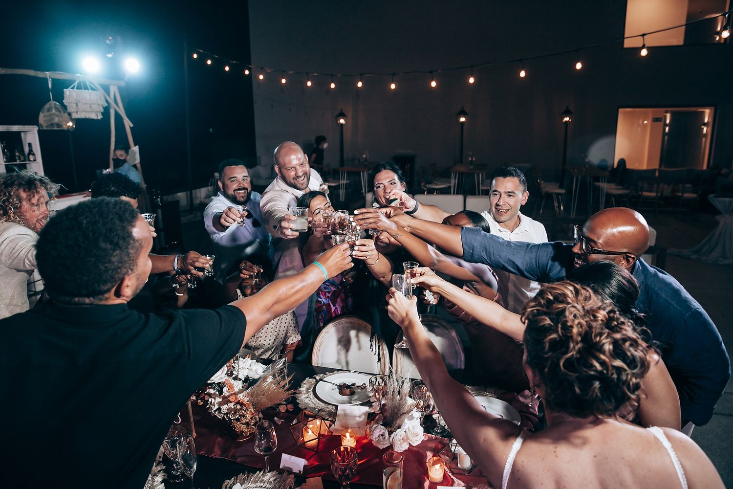 A group of people celebrating at a dimly lit party with string lights and raised drinks.