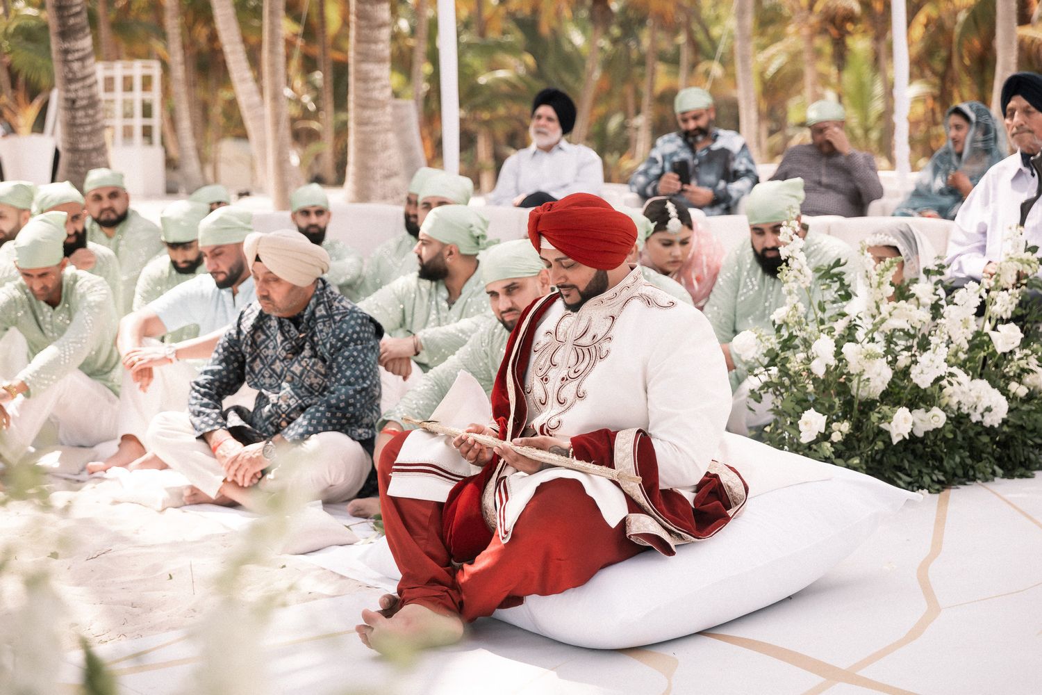 Sikh wedding ceremony outdoors with groom in red turban and white sherwani seated during Anand Karaj rituals.