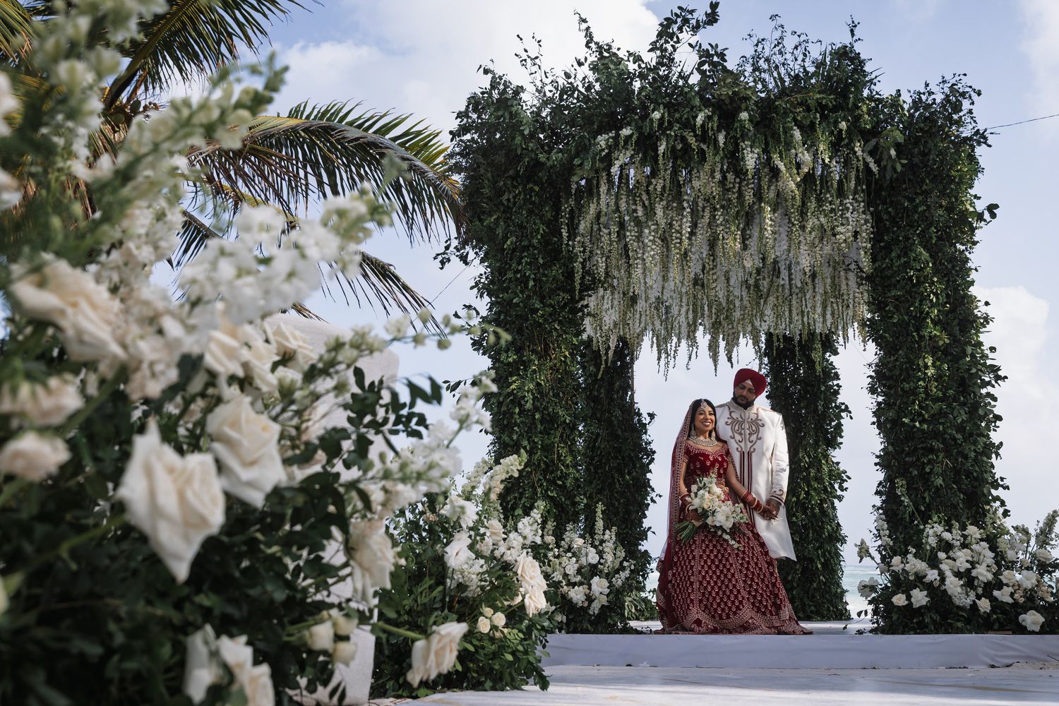 Bride in red lehenga and groom stand under lush greenery arch with white floral arrangements at outdoor wedding ceremony.