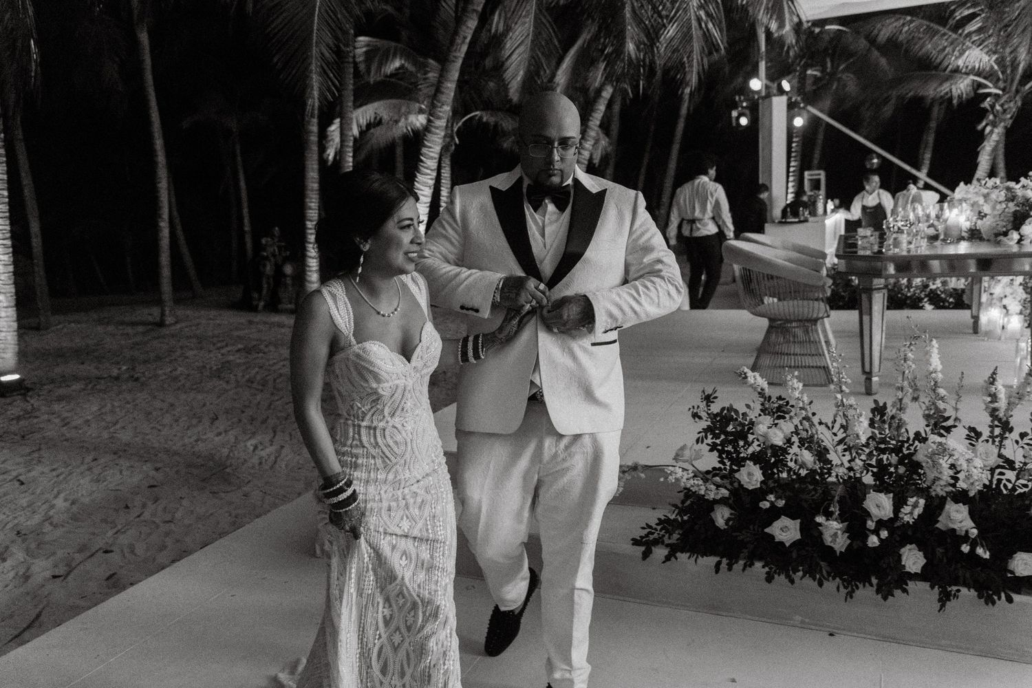 Black and white photo of bride in lace gown walking with escort at beach wedding.