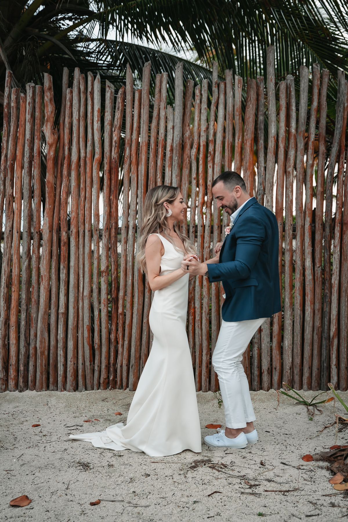 Bride and groom exchanging vows on sandy beach in front of rustic wooden fence with palm trees.