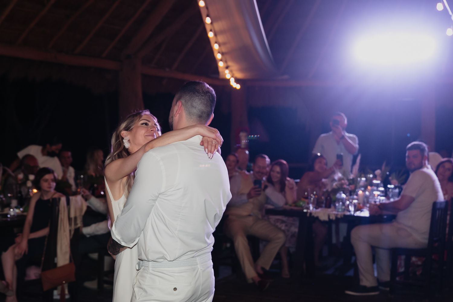 Bride and groom sharing romantic first dance under string lights at tropical wedding reception.