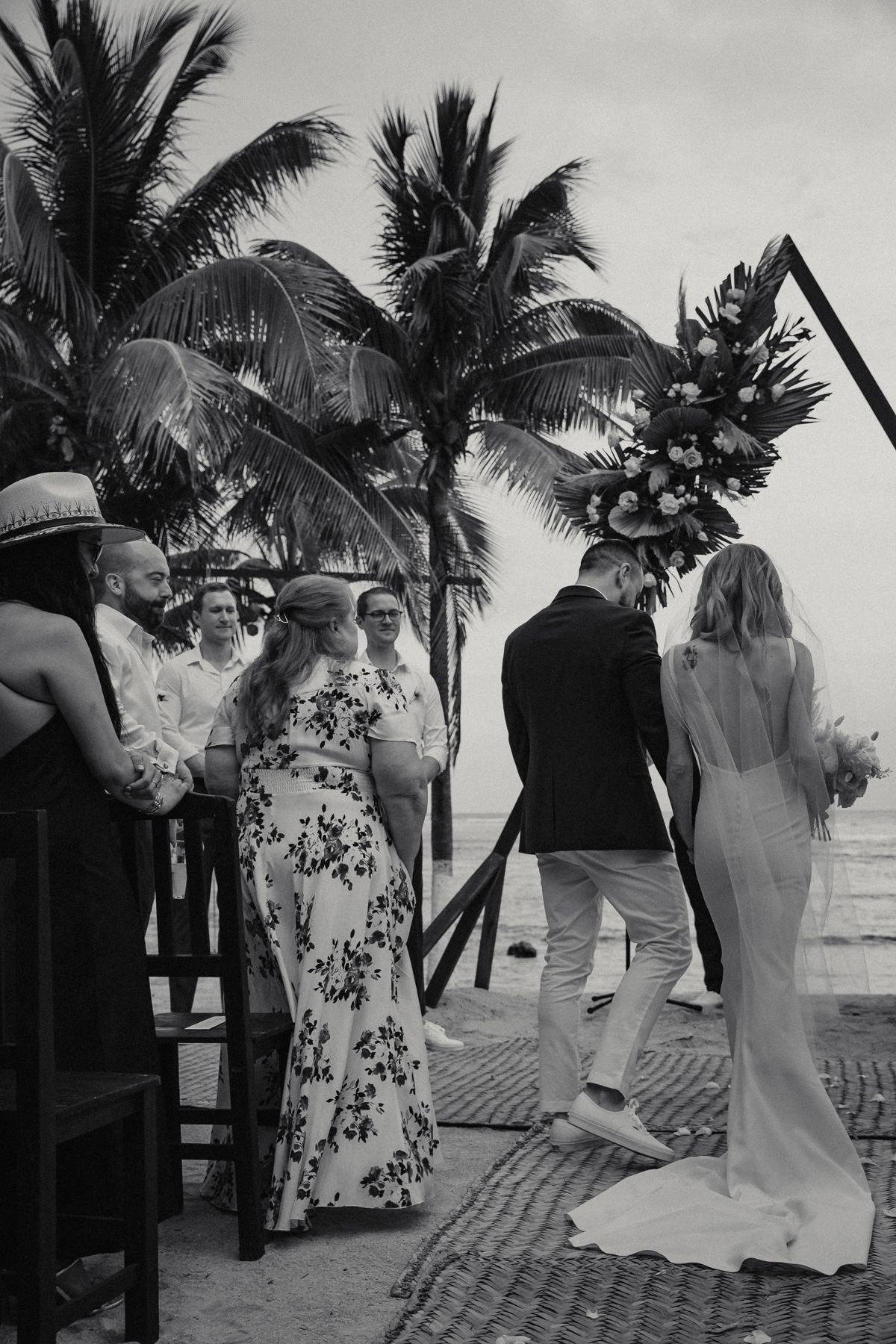 Bride and groom approach floral arch at tropical beach wedding with palm trees and guests watching.