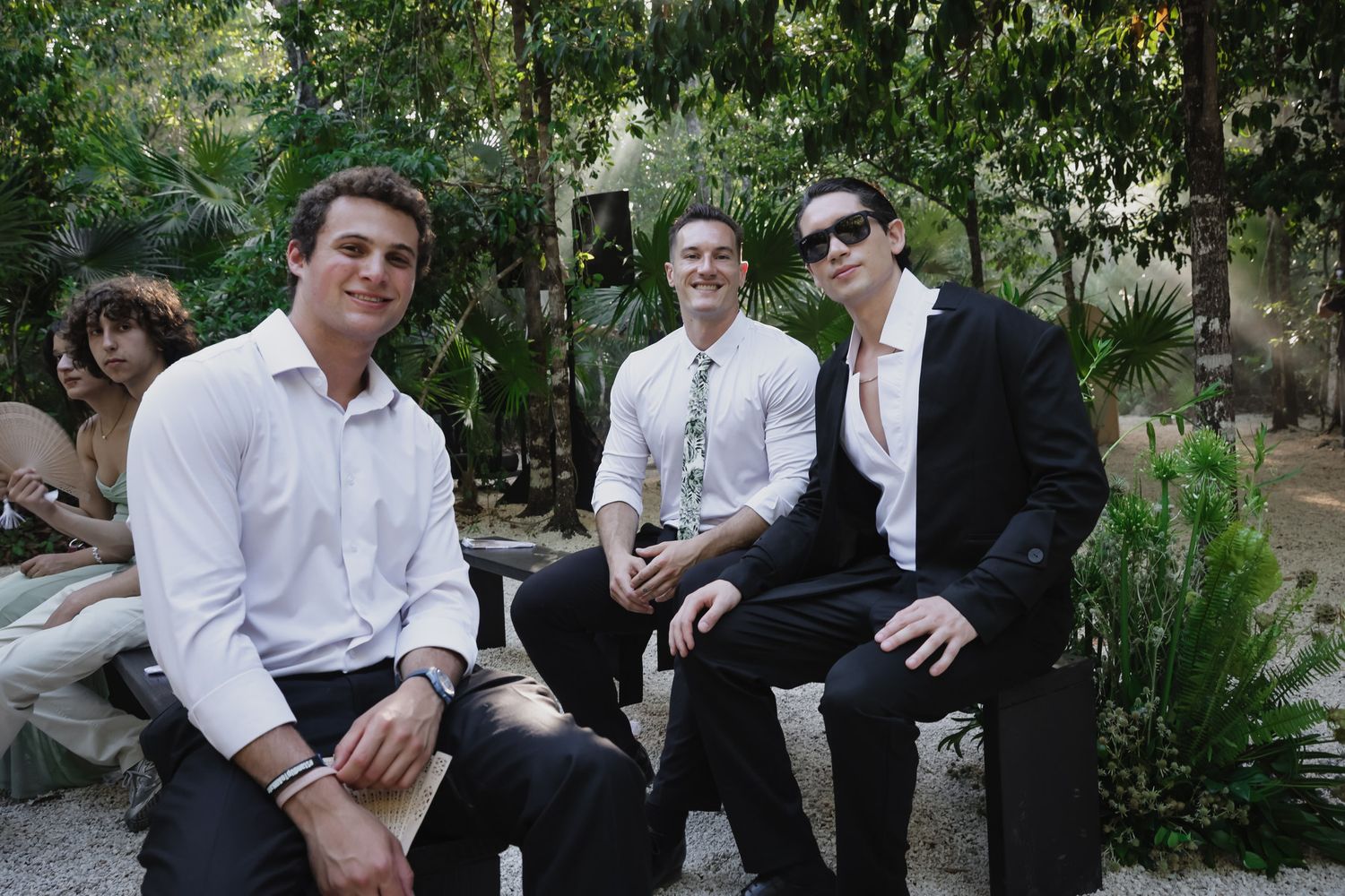 Three young men in formal attire smile while seated outdoors at a garden wedding venue.