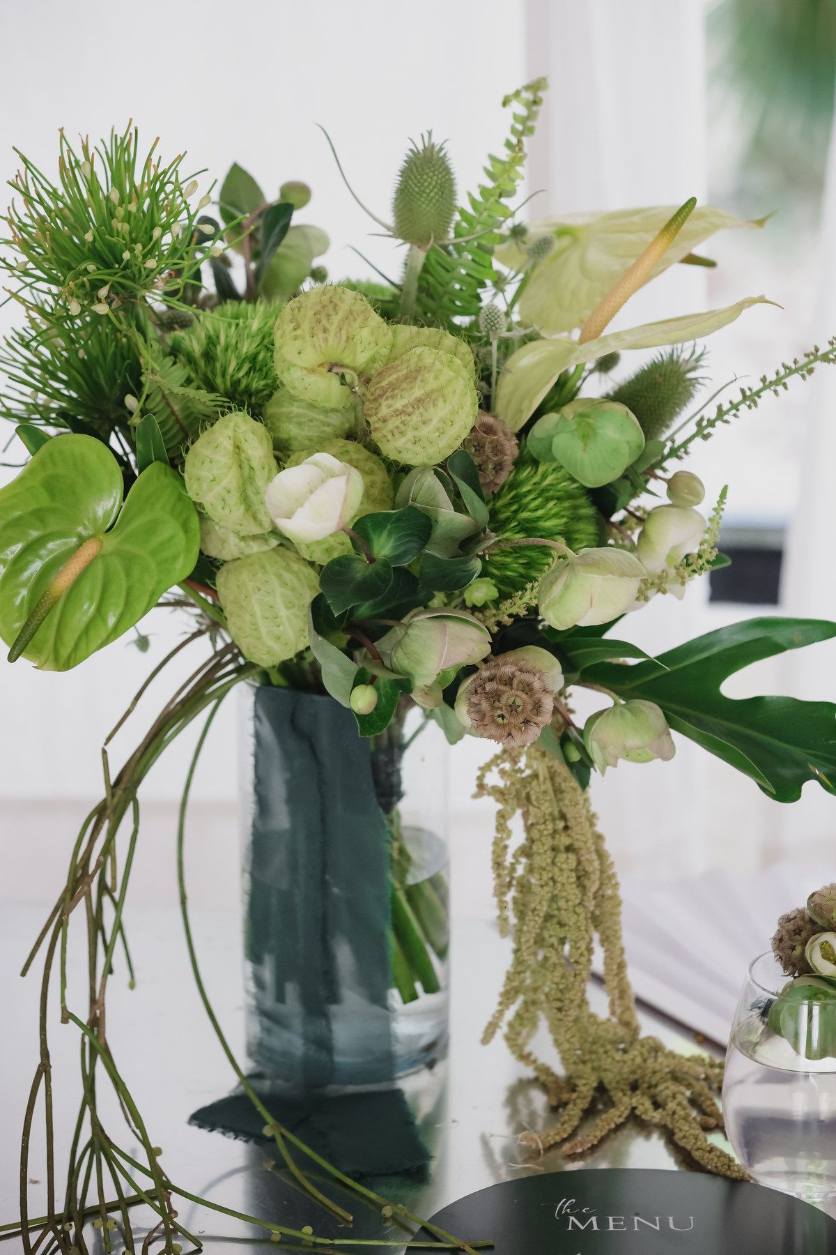 Lush green wedding floral arrangement in a glass vase featuring tropical leaves, white roses, and textured blooms.