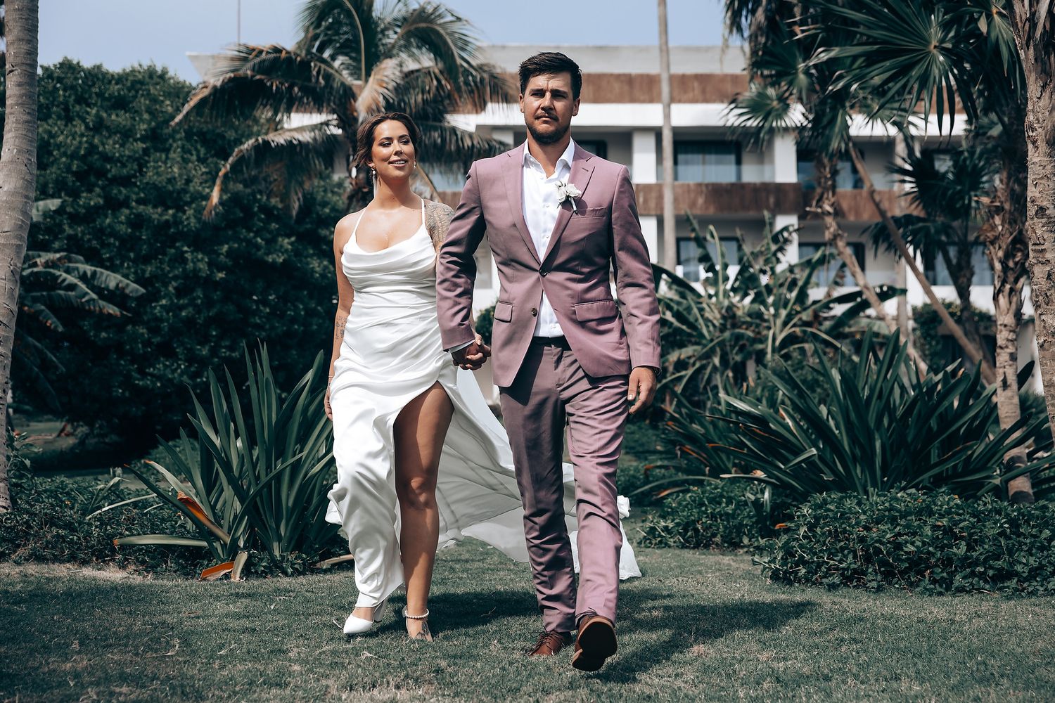 Wedding party walking along a tropical beach path lined with palm trees in coordinated pink and white attire.