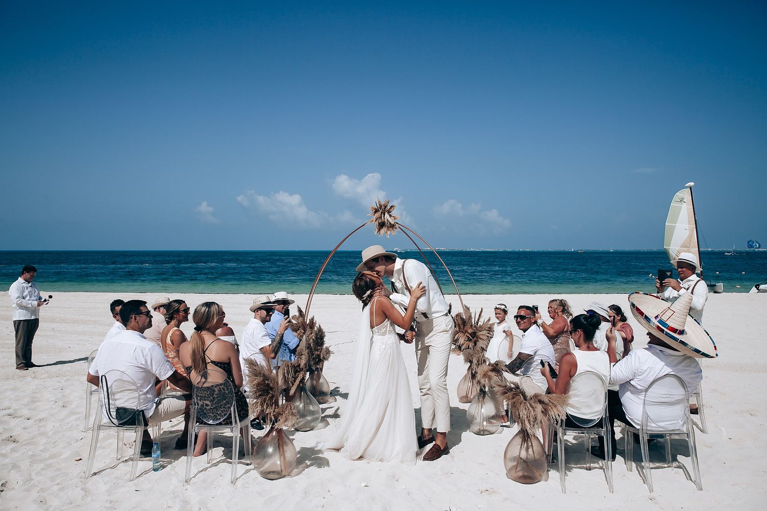 A panoramic view of a beach wedding setup with white chairs and guests gathered on a sunny tropical shoreline.
