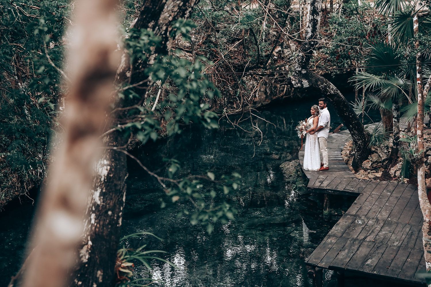 A romantic forest photoshoot featuring a couple in white clothing near trees and water.
