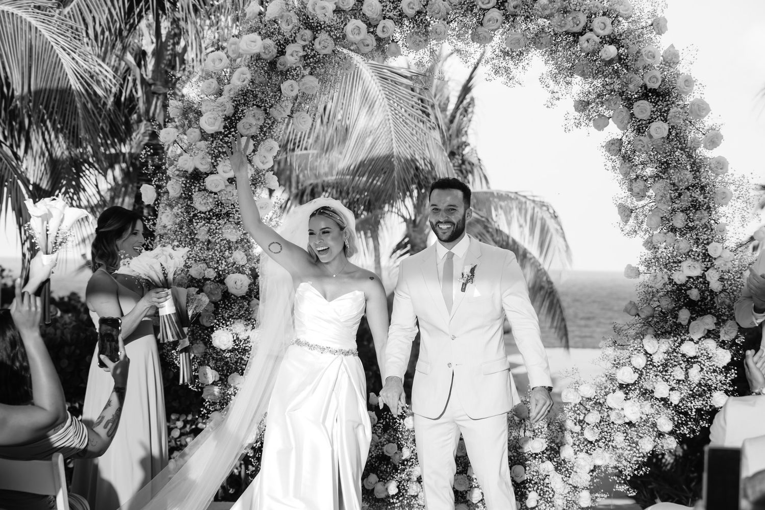 Black and white photo of newlyweds celebrating under floral arch with ocean view backdrop.
