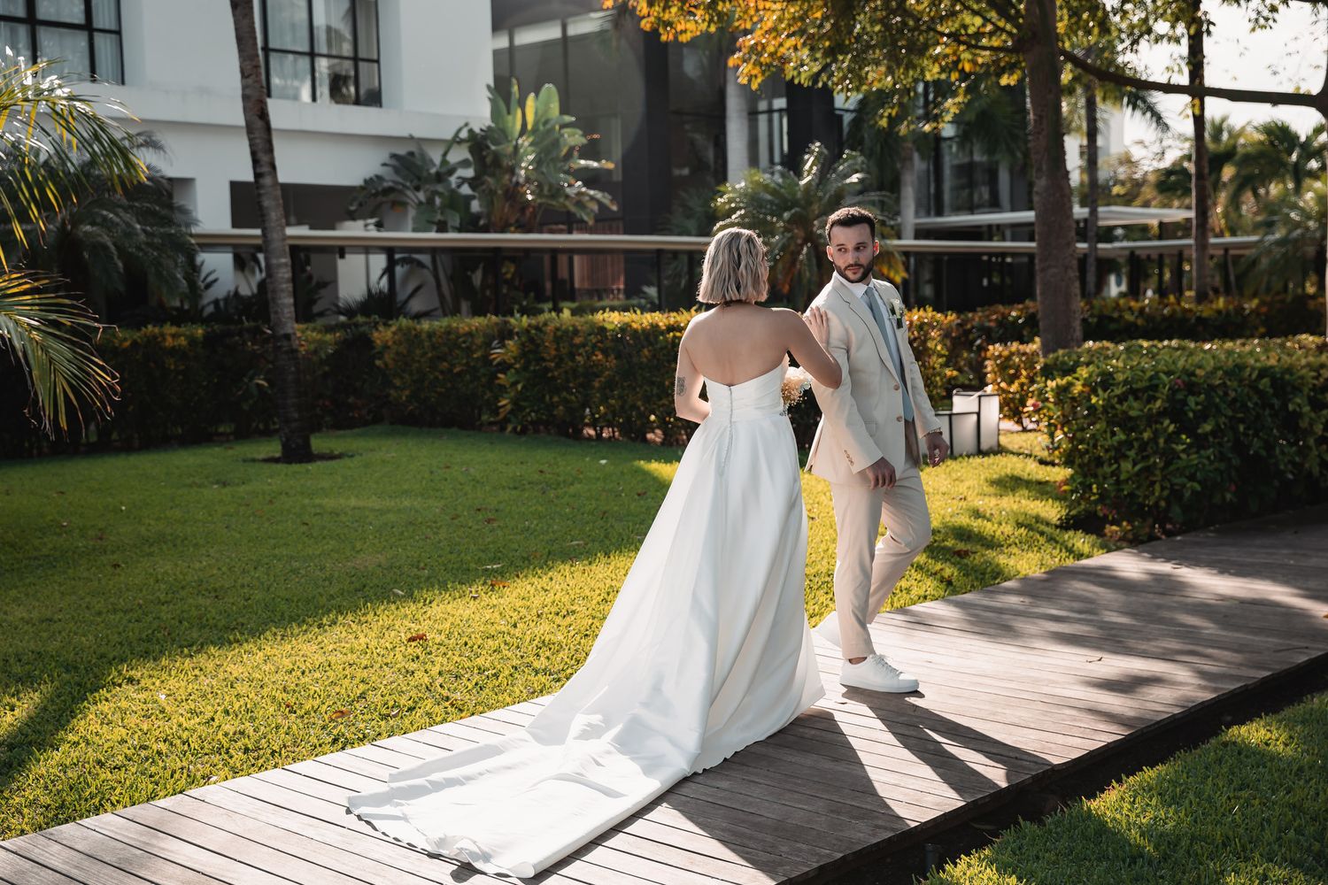 Bride and groom walking hand in hand on a wooden path through a lush green garden venue.
