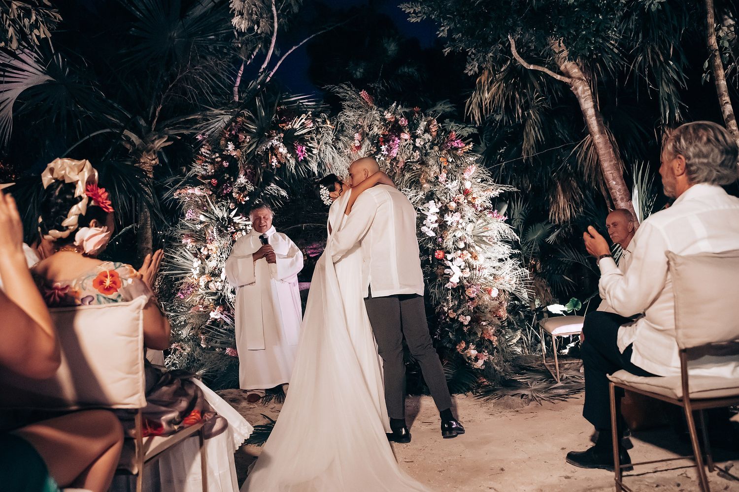 Wedding guests in white attire gather for an outdoor celebration under palm trees.
