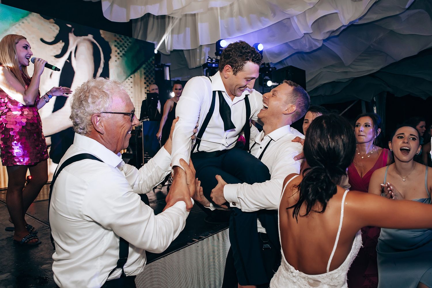 Wedding reception dance party with vibrant lighting and guests dancing under draped fabric decor.