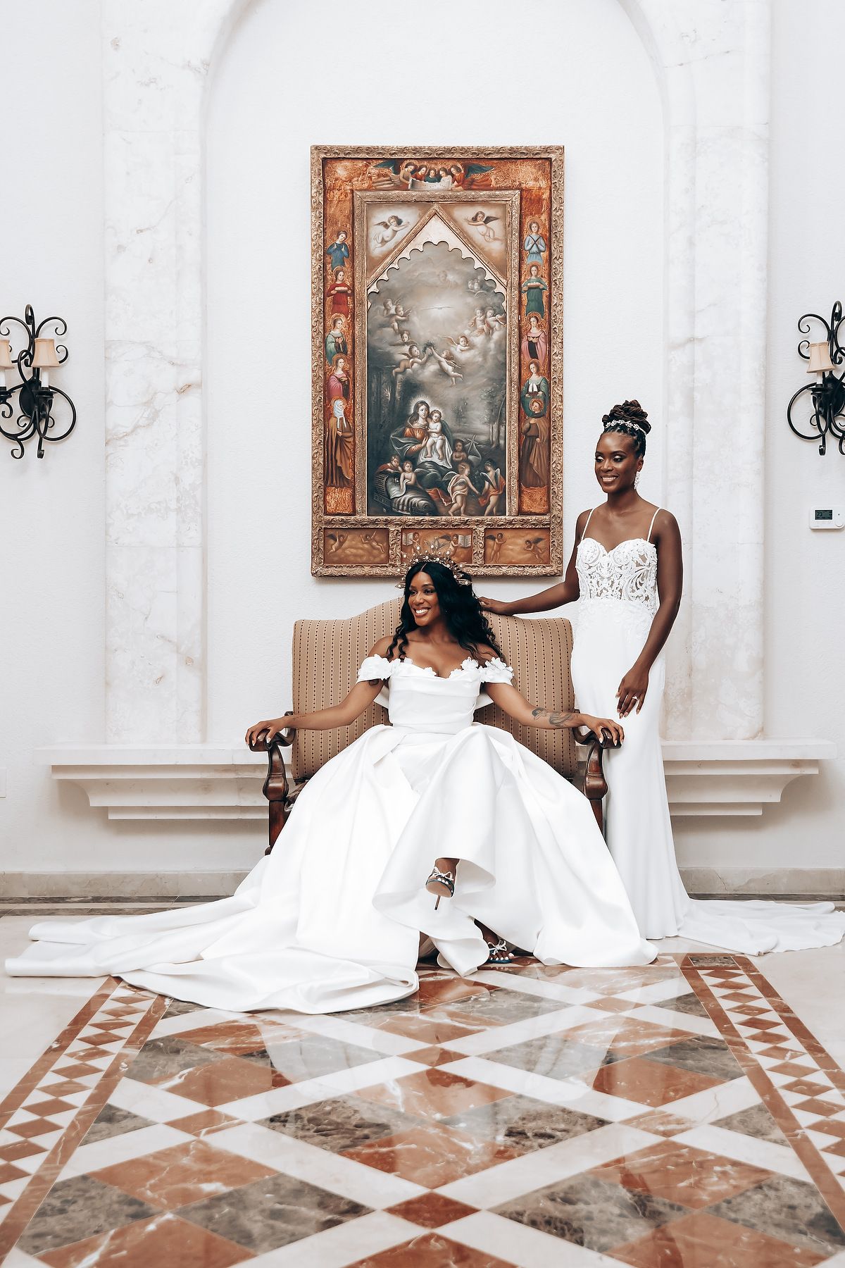 A regal wedding portrait in white gowns against ornate classical architecture and artwork.