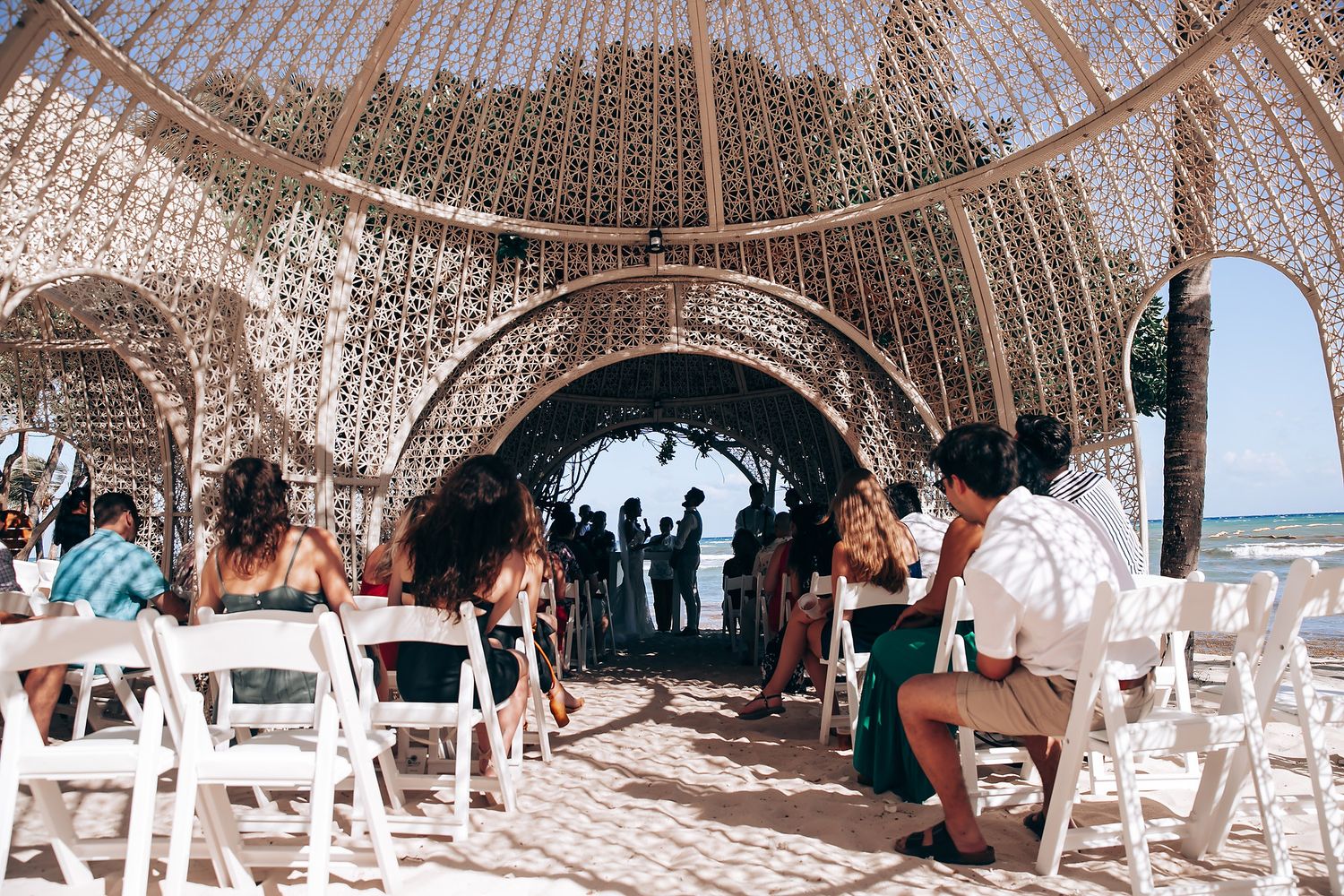 Wedding ceremony setup with white chairs under an elegant canopy structure on the beach.
