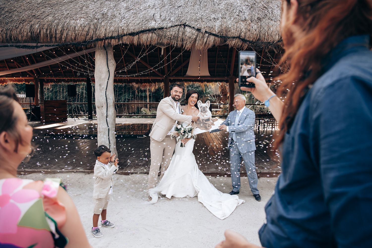 Wedding guests celebrate and dance during reception at an open-air beachside venue with thatched roof.