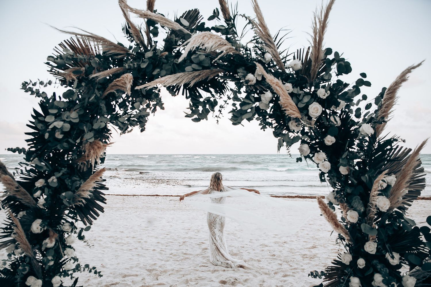 A decorative wedding arch with dark foliage and flowers frames a distant beach scene.