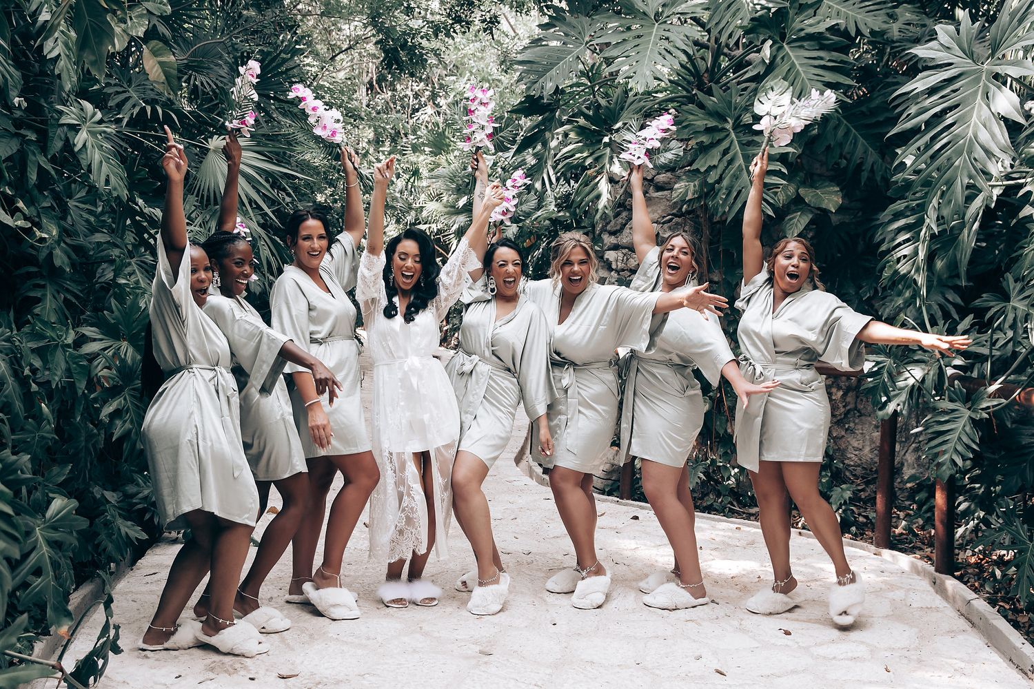 Wedding party in white robes poses playfully against lush tropical foliage.