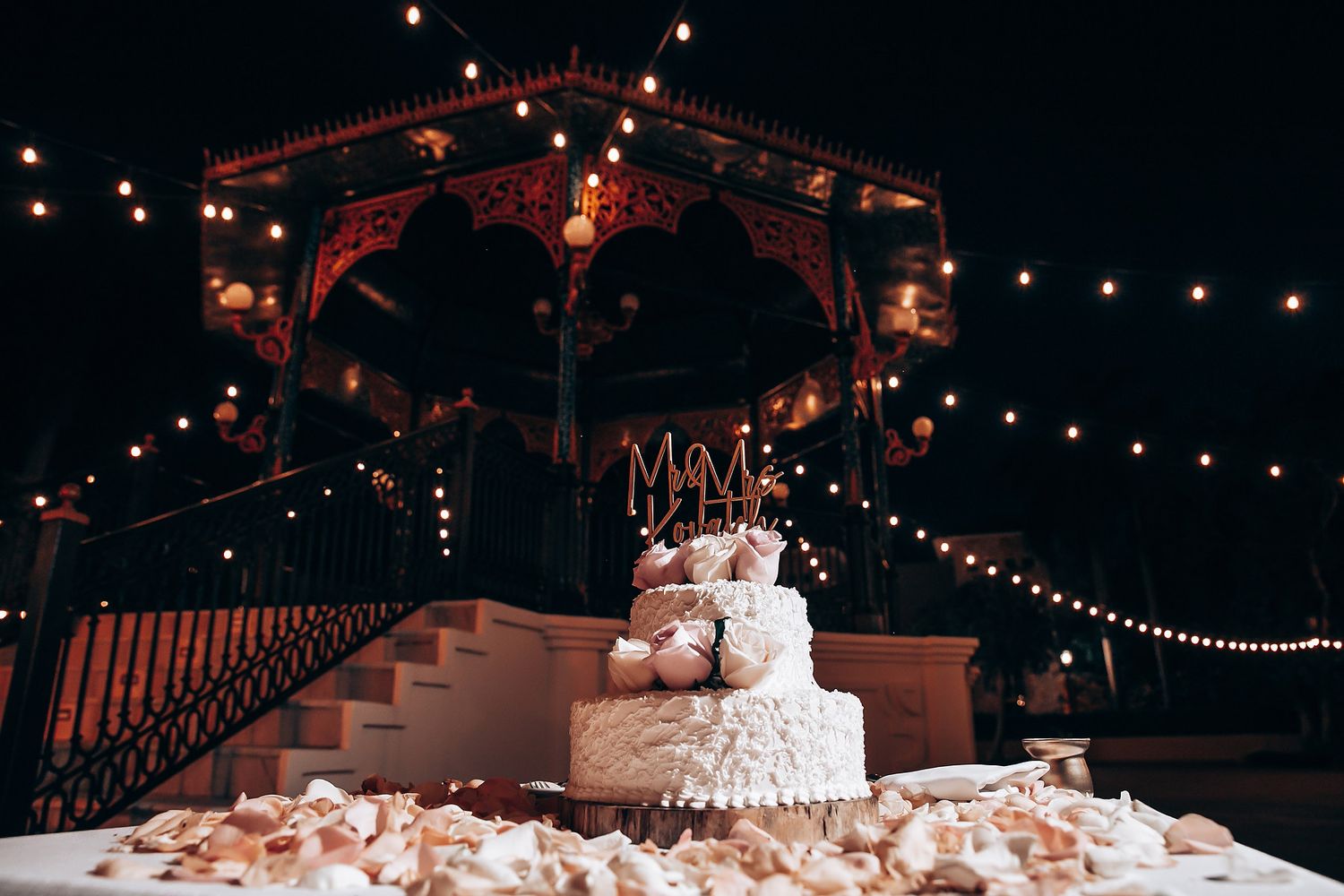 A tiered white wedding cake decorated with flowers sits under string lights at a rustic reception venue.
