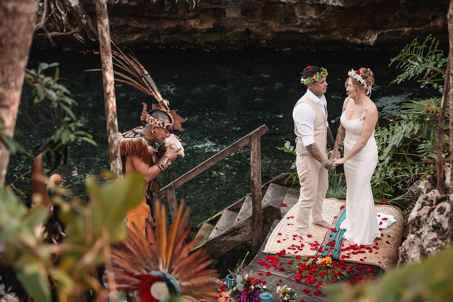 Couple exchanges vows at a cenote wedding ceremony with a Mayan officiant, rose petals, and lush jungle backdrop.
