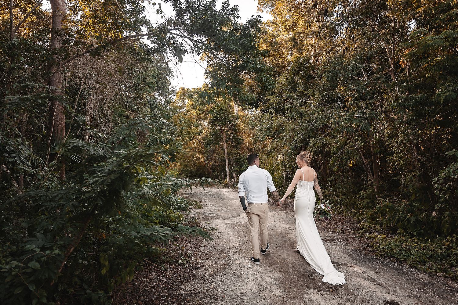 Newlyweds walk hand in hand down a scenic forest path during their outdoor wedding celebration.
