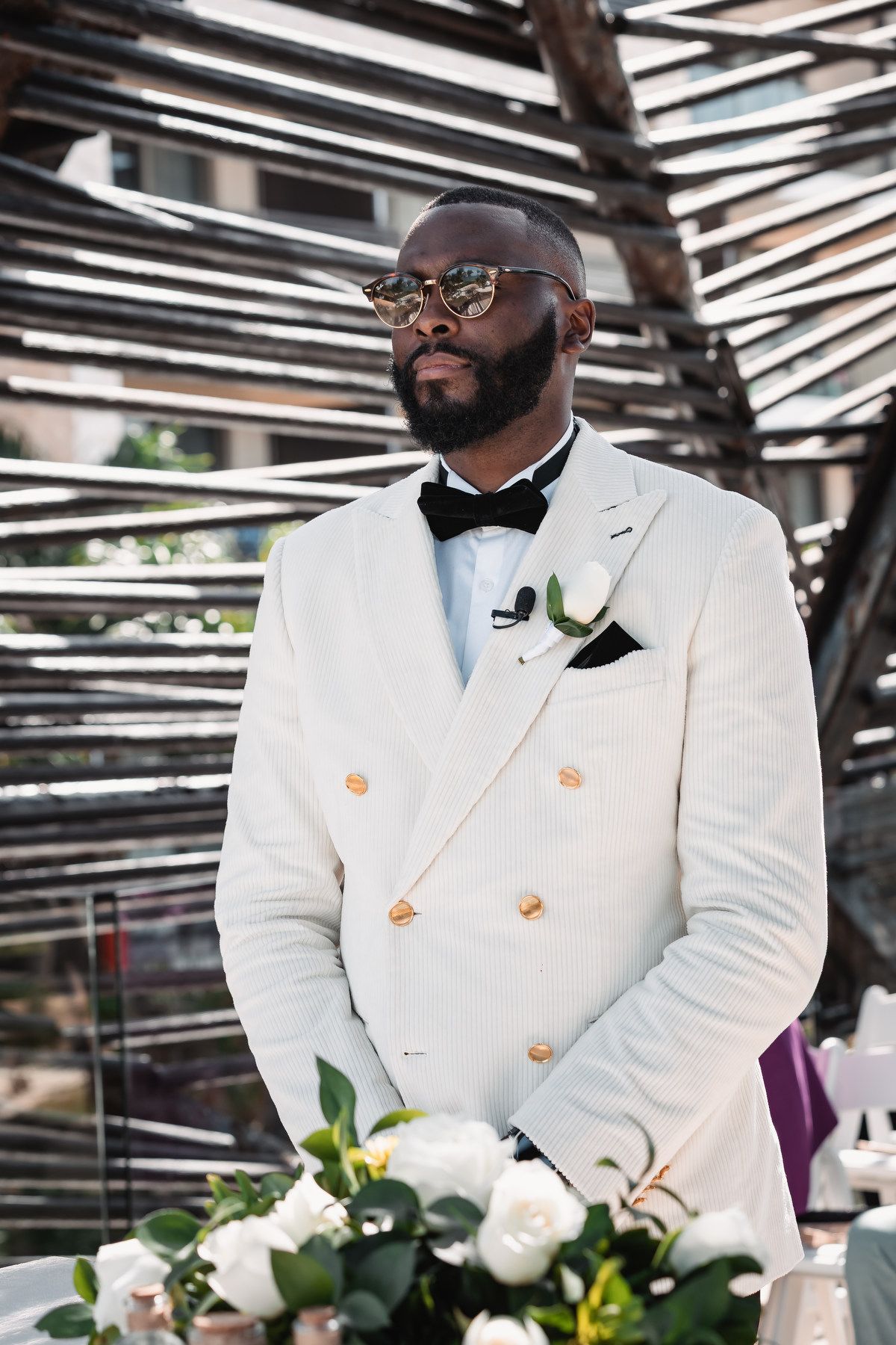Dapper Black groom in white double-breasted tuxedo with bow tie and boutonniere at outdoor wedding.