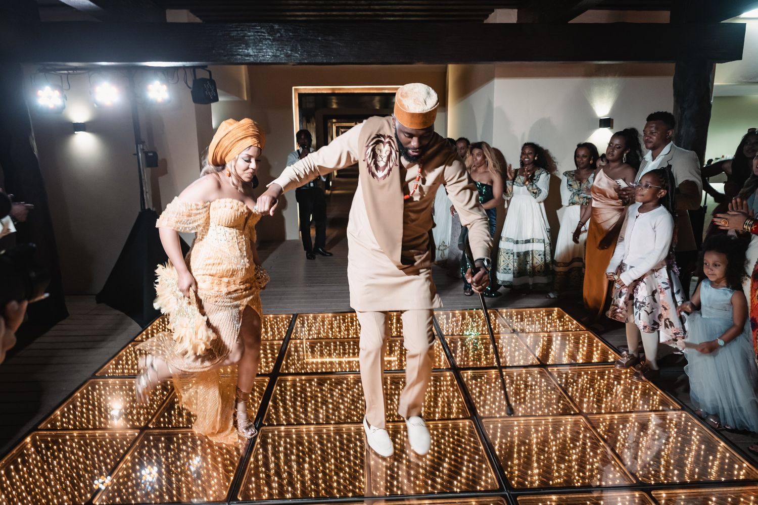 Couple dancing joyfully on illuminated dance floor at traditional African wedding celebration with guests watching.